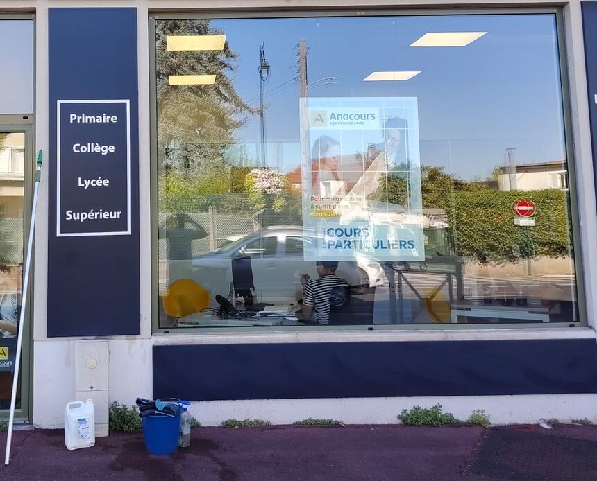 Grande vitrine d’une agence de soutien scolaire après le lavage, avec le seau, les raclettes, un bidon de produit et la perche posés au pied de la façade.